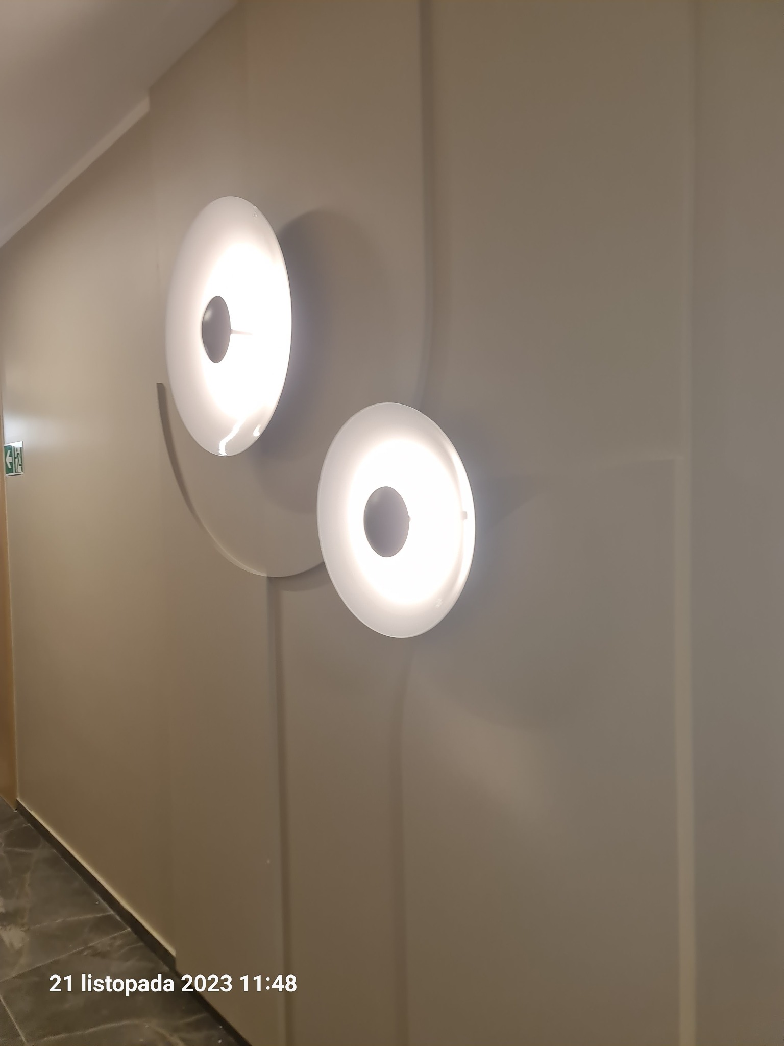 Dwa okrągłe, nowoczesne kinkiety ścienne z białym światłem, zamontowane na szarej ścianie z dekoracyjnymi panelami. Jeden kinkiet jest większy i umieszczony wyżej, tworząc asymetryczną kompozycję.