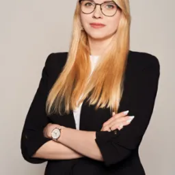 Portret kobiety w okularach, czarnej marynarce i z zegarkiem na ręku, stojącej na neutralnym tle z założonymi rękami.