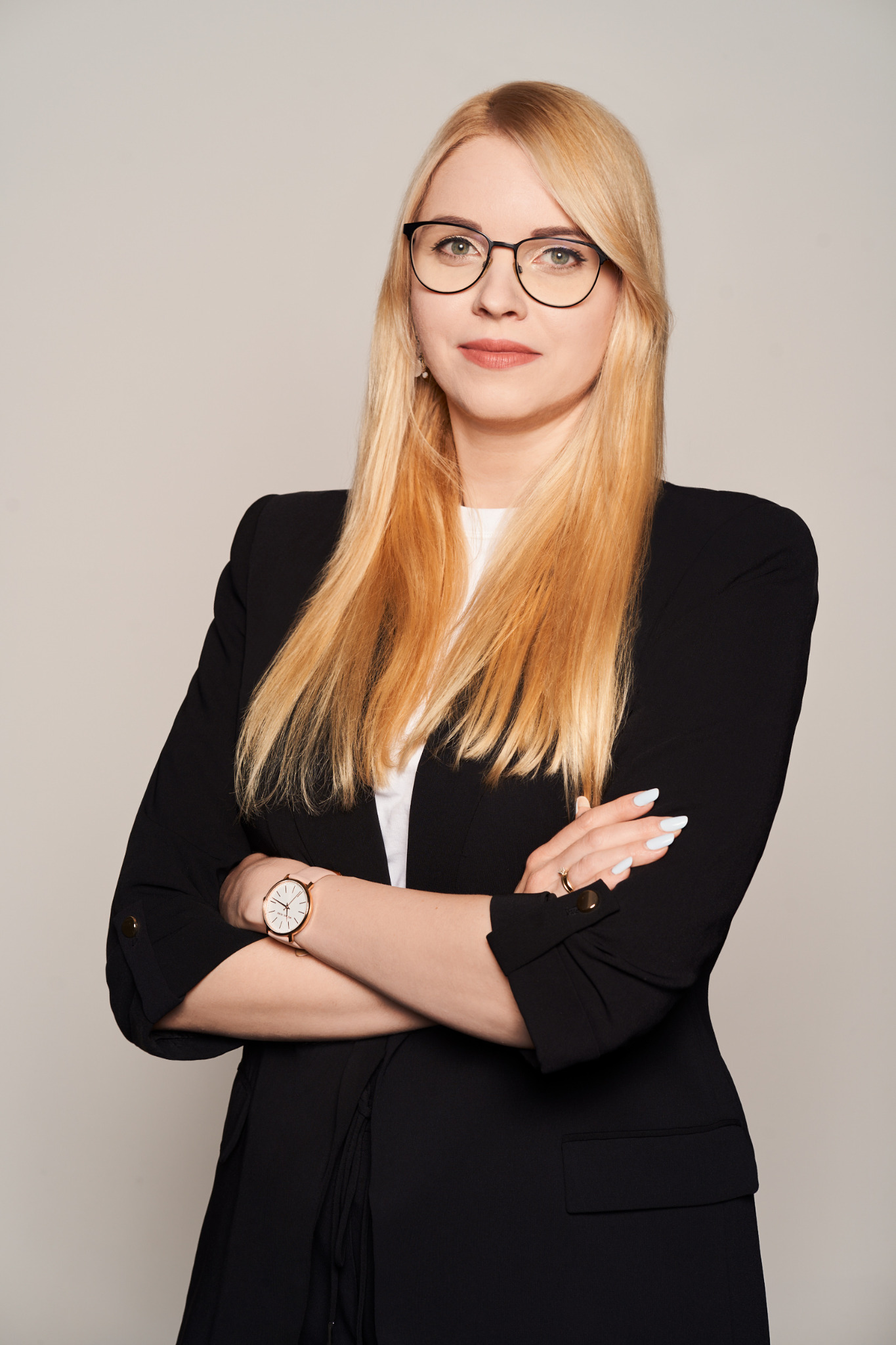 Portret kobiety w okularach, czarnej marynarce i z zegarkiem na ręku, stojącej na neutralnym tle z założonymi rękami.