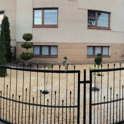Ogrody - Nowoczesny ogród przed domem z jasnym żwirem, czarnym metalowym ogrodzeniem i formowanymi drzewkami, widok z zewnątrz przez otwartą bramę.