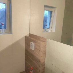 Mo-Kam bud - Montaż stelaża podtynkowego WC z obudową imitującą drewno, widoczne przyłącza kanalizacyjne i częściowo wykończone płytki ścienne w jasnym odcieniu, dwa okna z widokiem na zewnątrz.