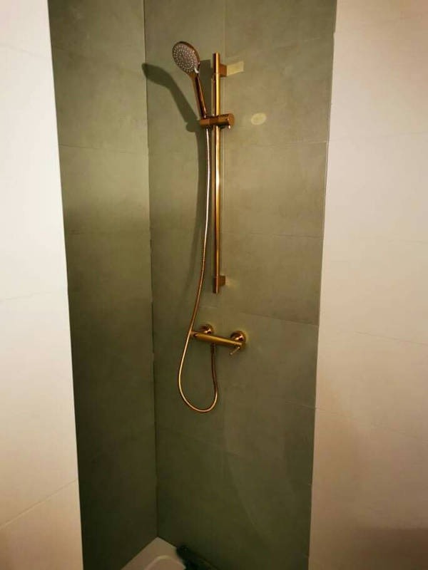 Nowoczesna kabina prysznicowa z deszczownicą i uchwytem w kolorze szczotkowanego złota na tle szarych płytek.