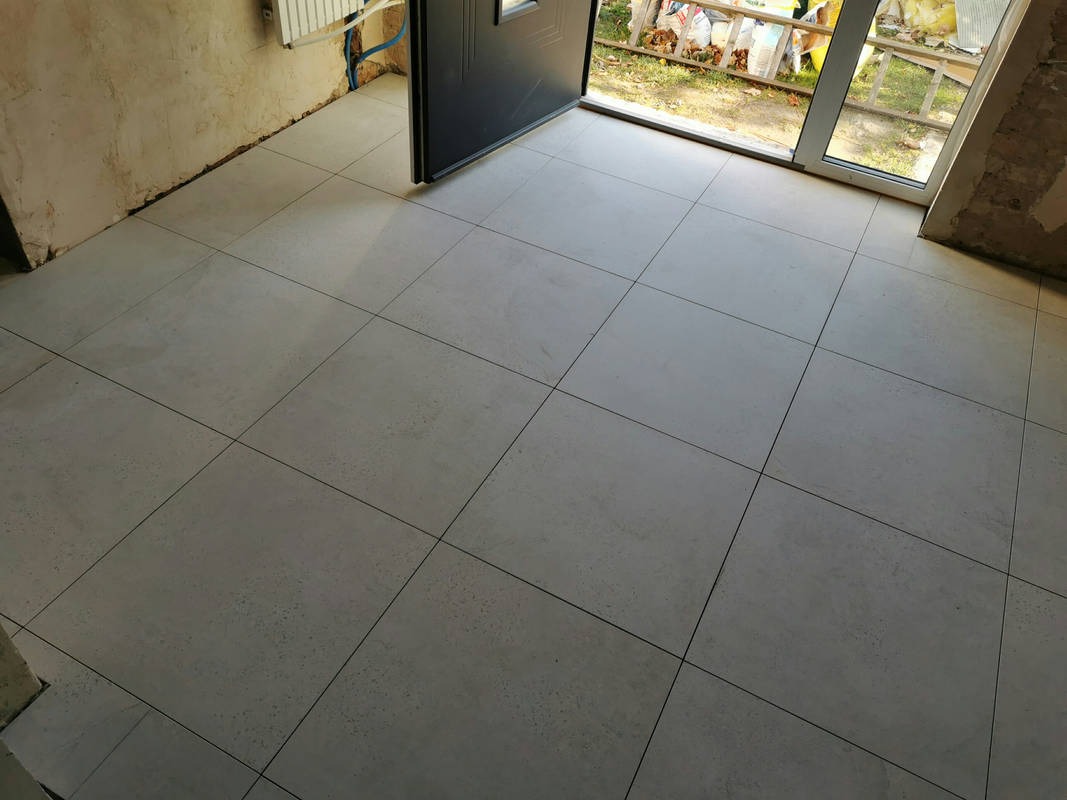 Ułożona podłoga z jasnych, kwadratowych płytek ceramicznych z ciemnymi fugami w pomieszczeniu w trakcie prac wykończeniowych, widoczne surowe ściany i otwarte drzwi wejściowe oraz okno z widokiem...