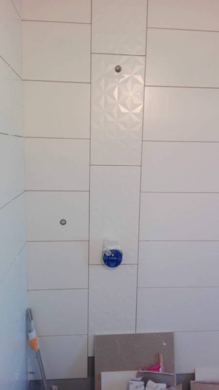 Białe, prostokątne płytki ceramiczne na ścianie łazienki, z pionowym pasem płytek z geometrycznym wzorem 3D, widoczne otwory na armaturę i zabezpieczone rury, fragment podłogi z resztkami...