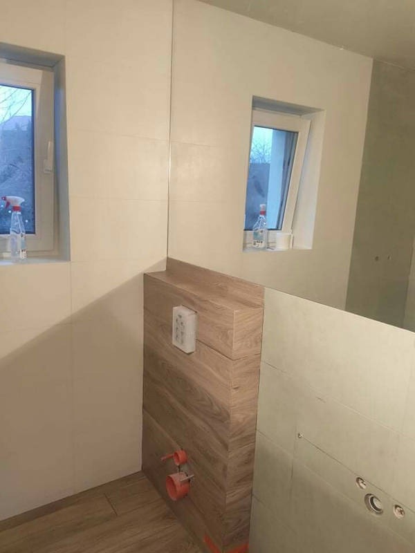 Montaż stelaża podtynkowego WC z obudową imitującą drewno, widoczne przyłącza kanalizacyjne i częściowo wykończone płytki ścienne w jasnym odcieniu, dwa okna z widokiem na zewnątrz.