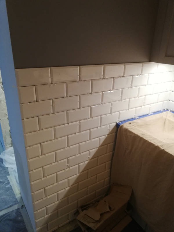 Świeżo położone białe, prostokątne płytki typu metro na ścianie, z widocznymi szarymi fugami, górna część ściany pomalowana na ciemnoszaro, widoczne zabezpieczenie folią malarską.