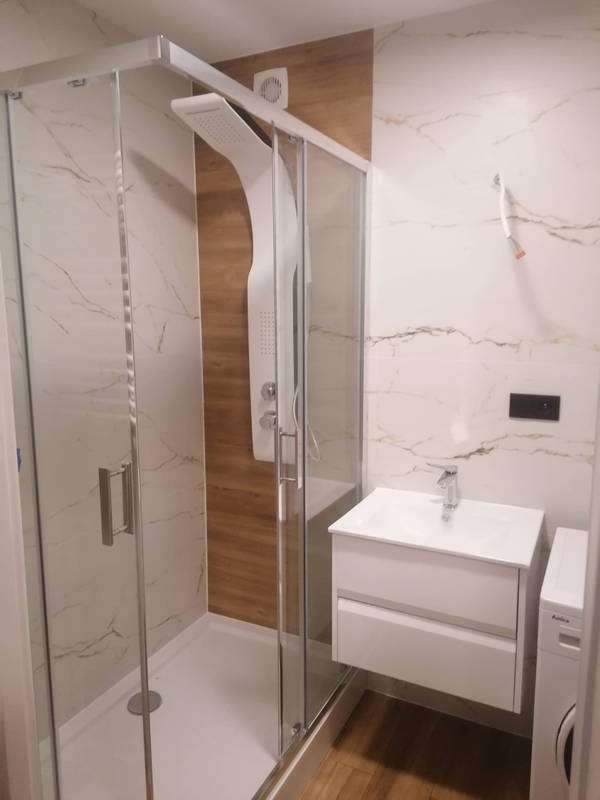 Nowoczesna łazienka z kabiną prysznicową typu walk-in z panelem natryskowym, białą umywalką z szafką i pralką obok, wykończona płytkami imitującymi marmur i drewno.