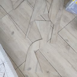 RemontSerwis - Fragmenty paneli podłogowych, przycięte do niestandardowych kształtów, leżą na podłodze z ułożonych paneli w odcieniach beżu i szarości, obok znajduje się worek kleju i fragment dekoracyjnej listwy...