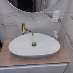 RemontSerwis - Biała, owalna umywalka nablatowa z złotą baterią i korkiem, stojąca na szafce z blatem imitującym drewno, na tle marmurowych płytek i okrągłego lustra z widocznym odbiciem osoby.