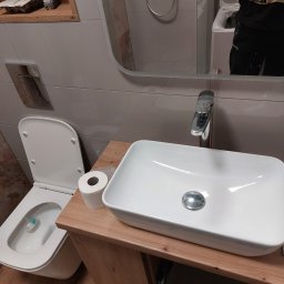 RemontSerwis - Nowoczesna łazienka z białą umywalką nablatową na drewnianej szafce, wisząca toaleta oraz lustro, w którym odbija się kabina prysznicowa i pralka.