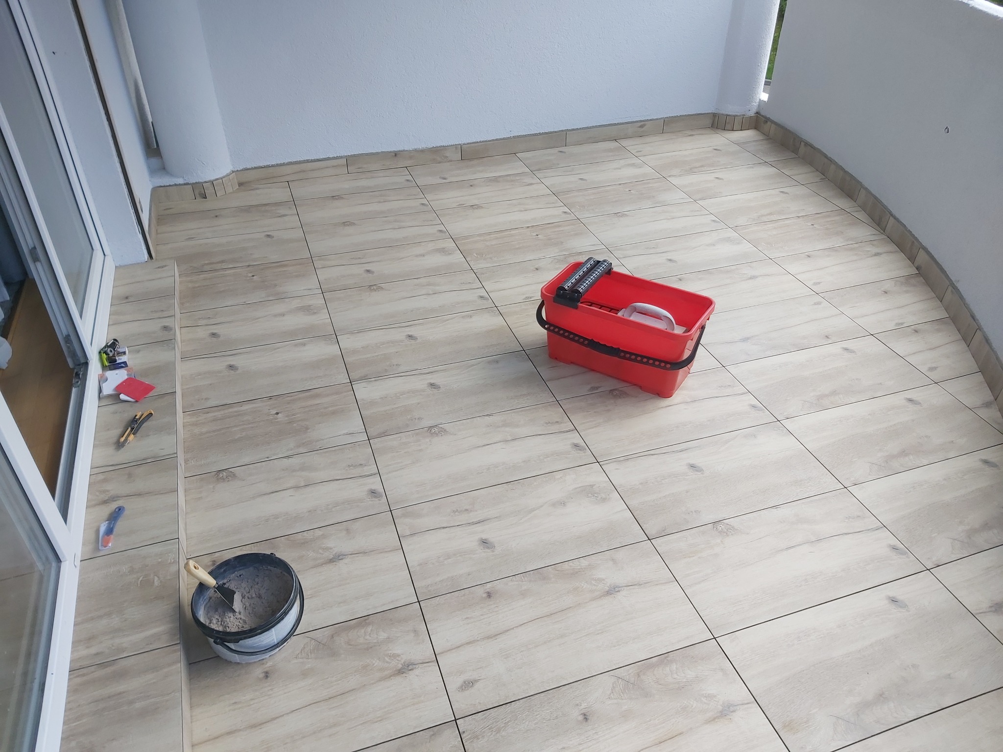 Balkon wyłożony płytkami imitującymi drewno, widoczny kubeł z zaprawą, szpachelka, czerwone wiadro i narzędzia do cięcia płytek.