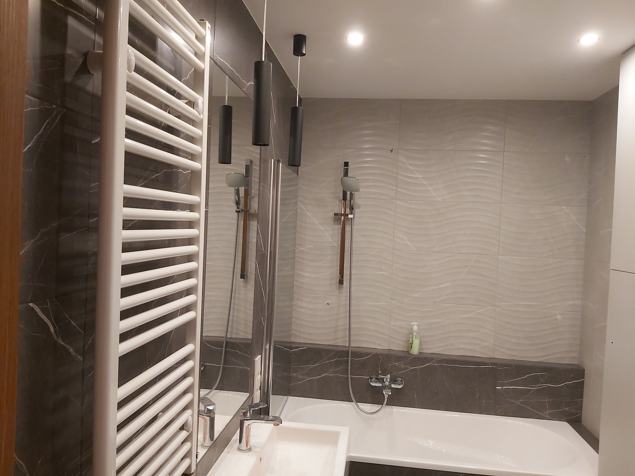 Nowoczesna łazienka z białym grzejnikiem drabinkowym, umywalką wpuszczaną w blat z szarego marmuru, wanną i płytkami imitującymi fale na ścianie.