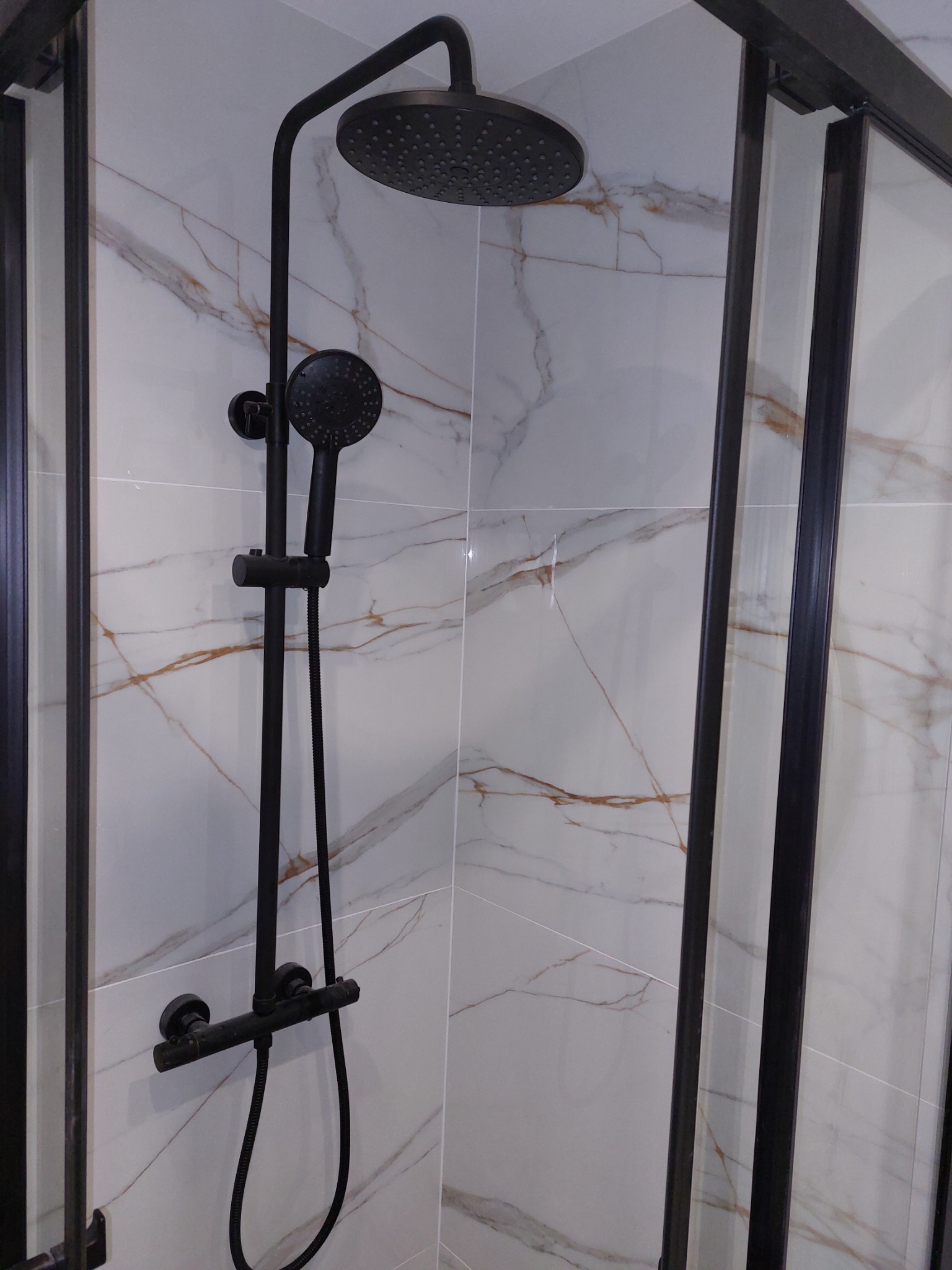 Czarny zestaw prysznicowy z deszczownicą i baterią termostatyczną na tle białych płytek imitujących marmur w kabinie prysznicowej z czarnymi profilami.