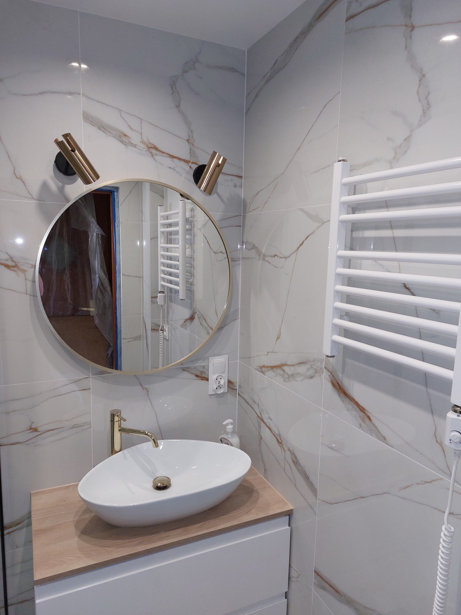 Nowoczesna łazienka z owalną umywalką nablatową, złotą armaturą, okrągłym lustrem podświetlonym złotymi lampami i grzejnikiem drabinkowym na ścianie wyłożonej marmurkowymi płytkami.