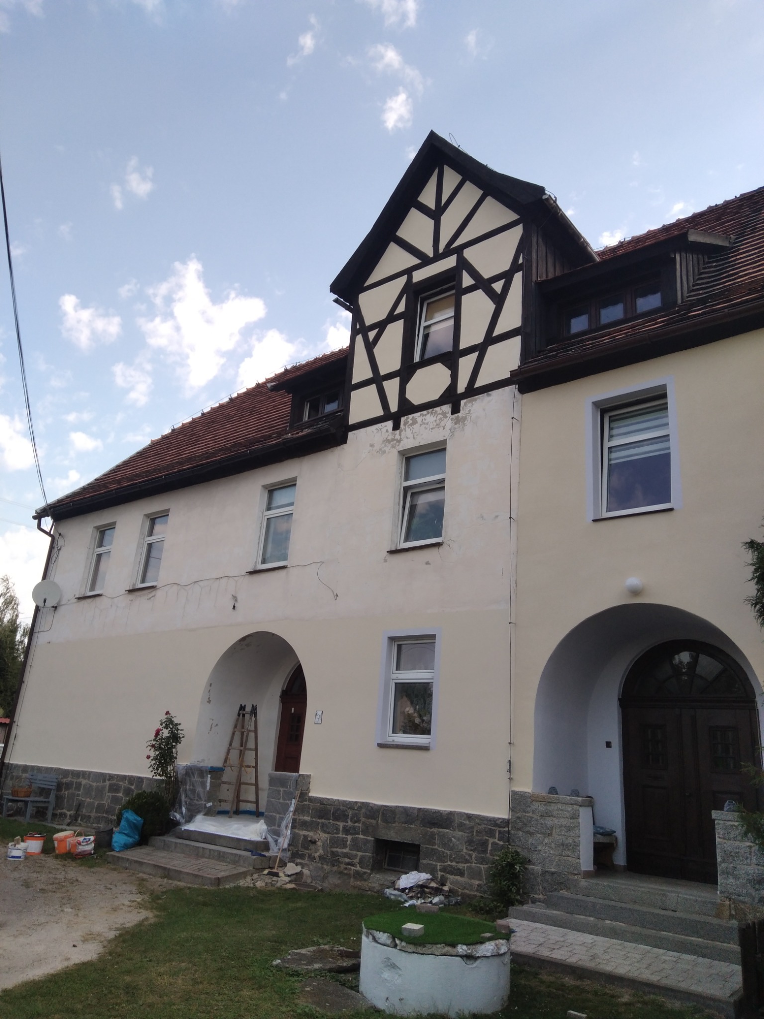 Dwupiętrowy dom z jasną elewacją w trakcie prac, widoczne pędzle, drabina i wiaderka z farbą przy wejściu.