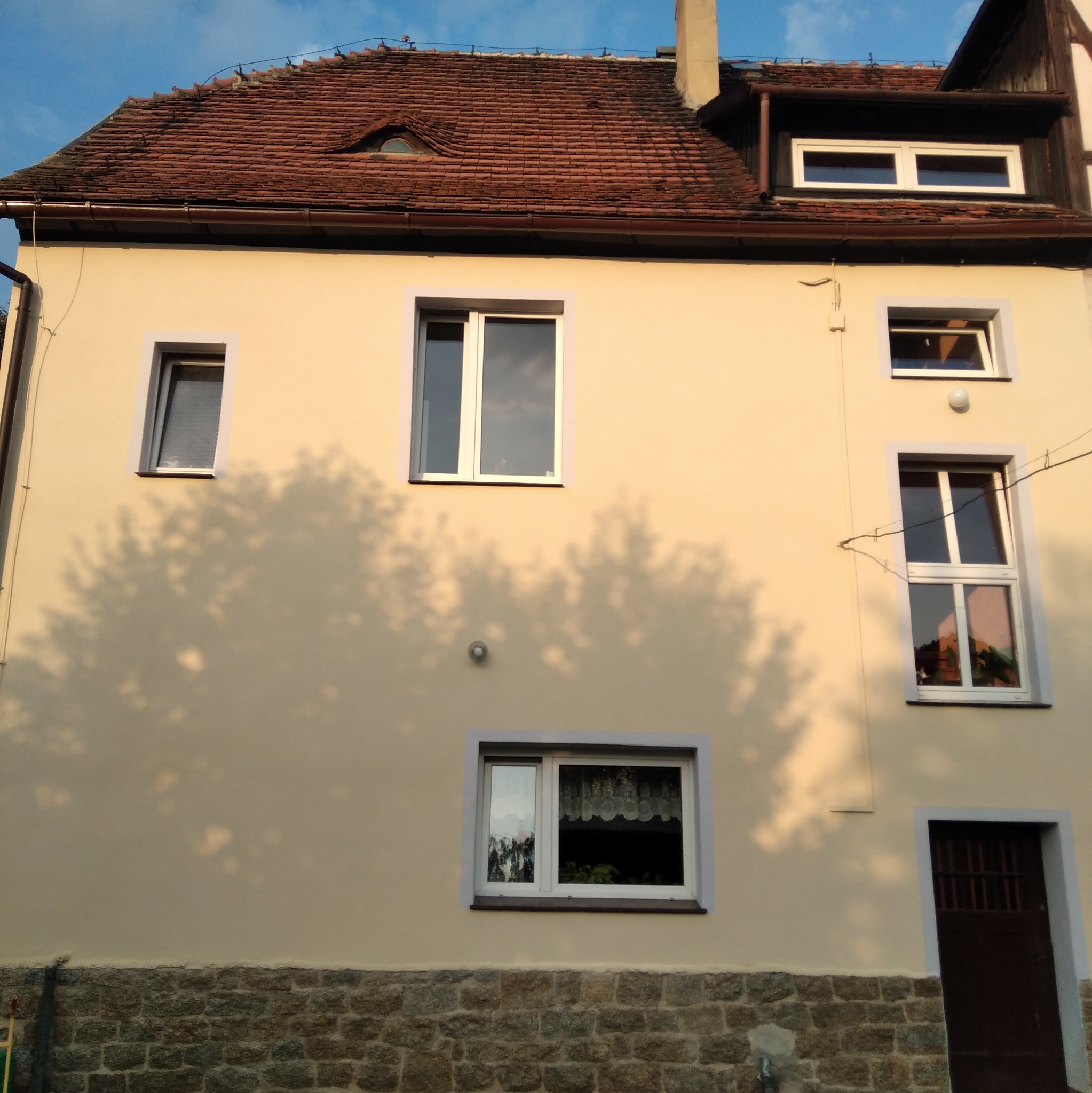 Odnowiona elewacja budynku z beżową farbą, widoczne okna z białymi ramami i kamienny cokół, cień drzewa na ścianie.