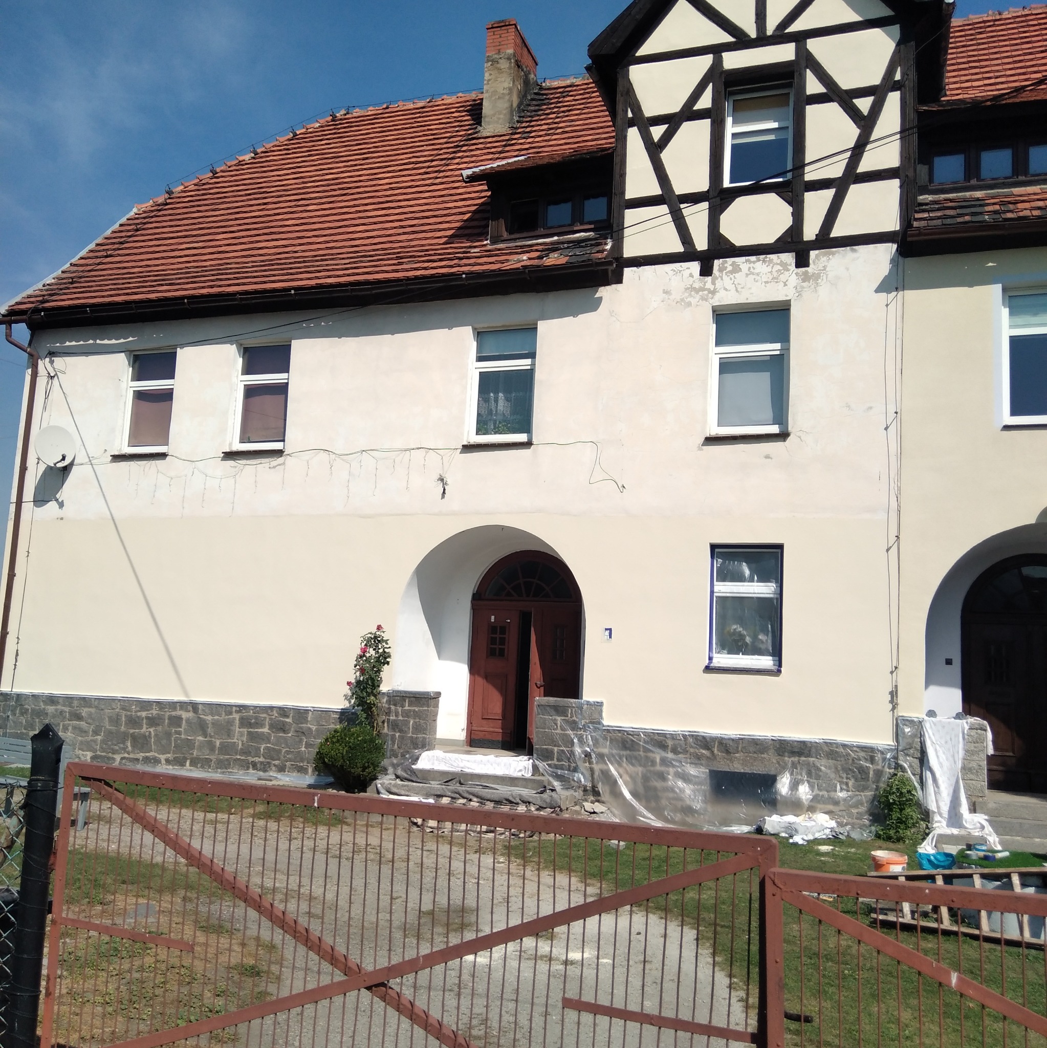 Odnowiona elewacja dwukondygnacyjnego domu z czerwonym dachem, widoczne rusztowania i zabezpieczenia przed malowaniem, brama wjazdowa na pierwszym planie.