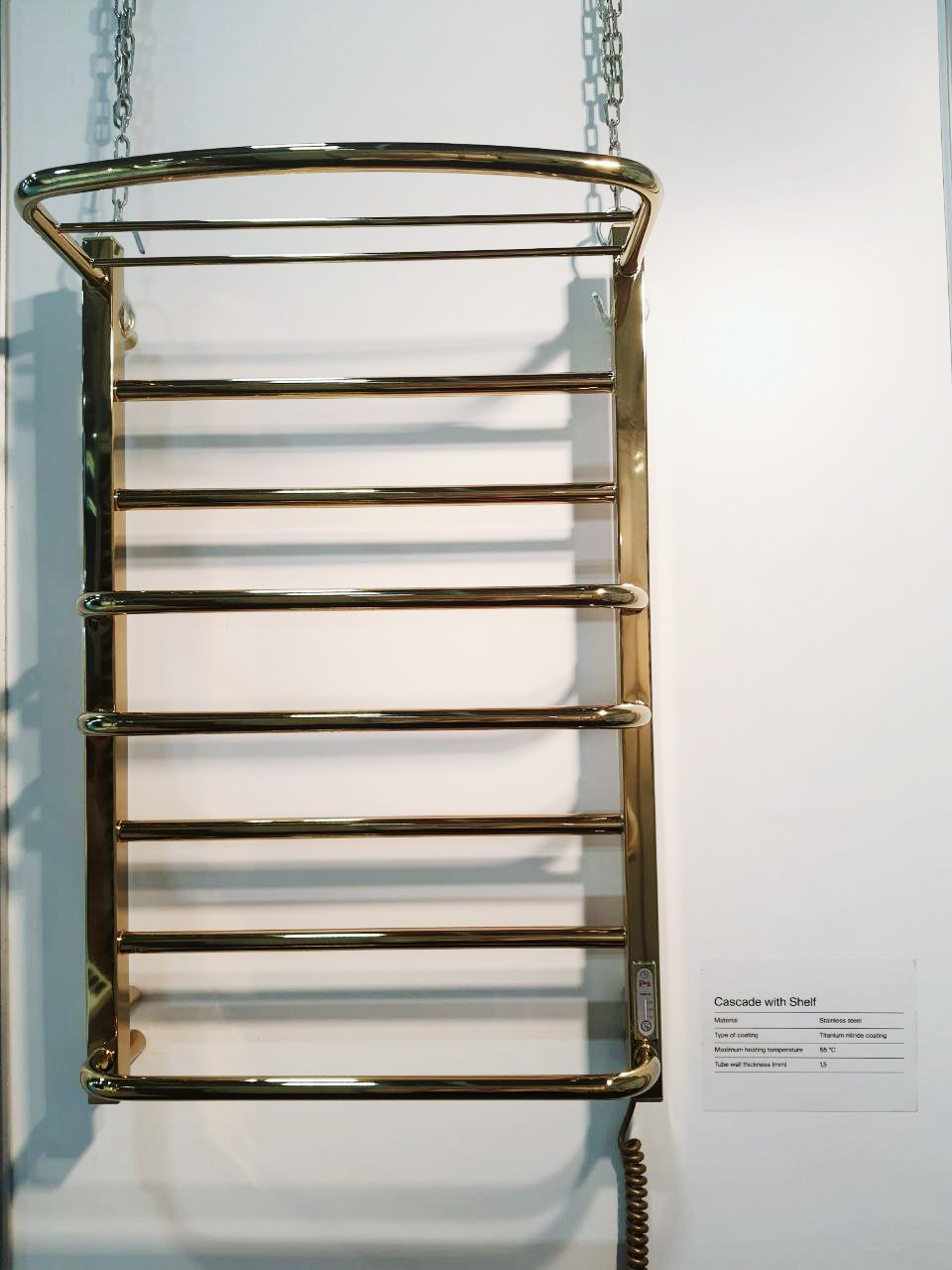 Złoty, elektryczny grzejnik łazienkowy z półką, wiszący na łańcuchach, z widocznym przewodem zasilającym i panelem sterowania, prezentowany na tle białej ściany z kartą informacyjną.