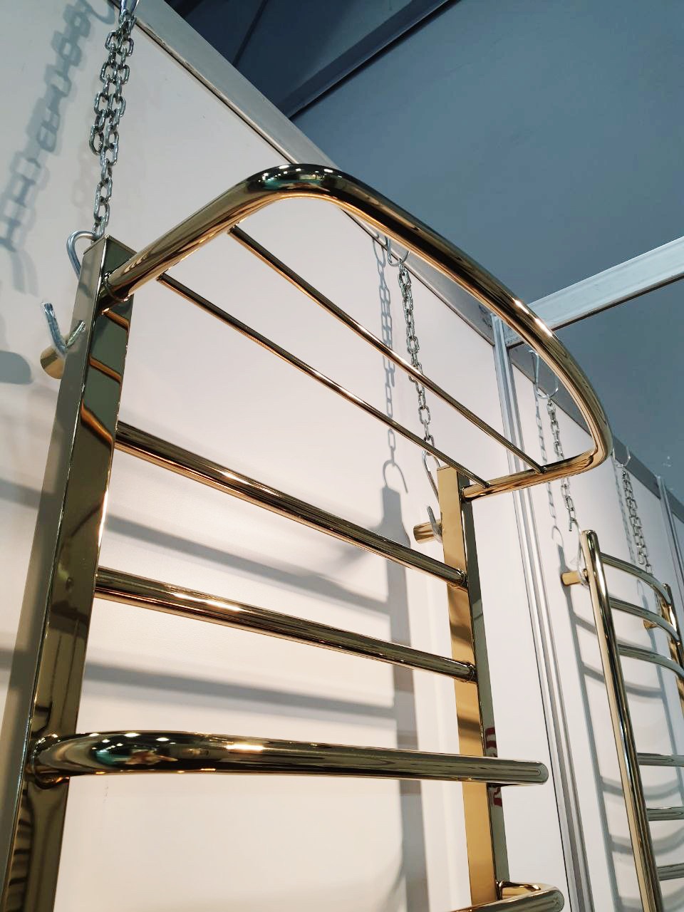 Błyszczący, złoty grzejnik drabinkowy łazienkowy, zawieszony na łańcuchach, z półką na ręczniki i widoczną częścią instalacji w tle.