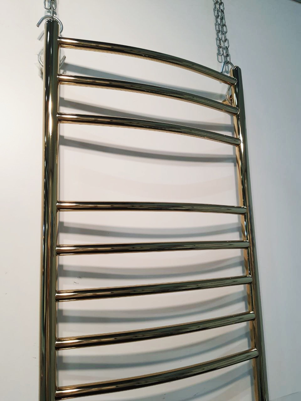 Błyszczący, złoty grzejnik łazienkowy drabinkowy, zawieszony na łańcuchu, prezentowany na tle białej ściany w Warszawie. Widoczne odbicia światła podkreślają luksusowy charakter.