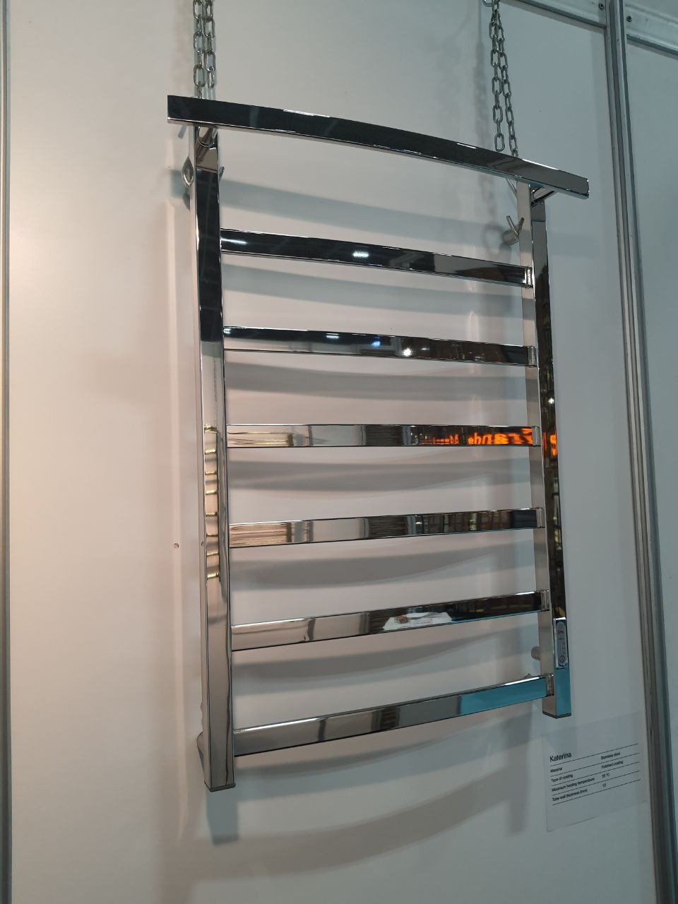 Błyszczący, chromowany grzejnik łazienkowy drabinkowy, wiszący na łańcuchach, z widocznym panelem sterowania po prawej stronie i specyfikacją techniczną na naklejce.