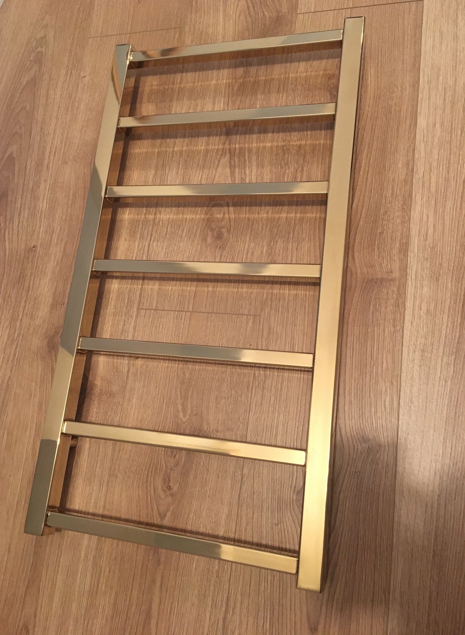 Złoty, błyszczący grzejnik łazienkowy drabinkowy z kwadratowymi rurami, leżący na podłodze z paneli imitujących drewno.