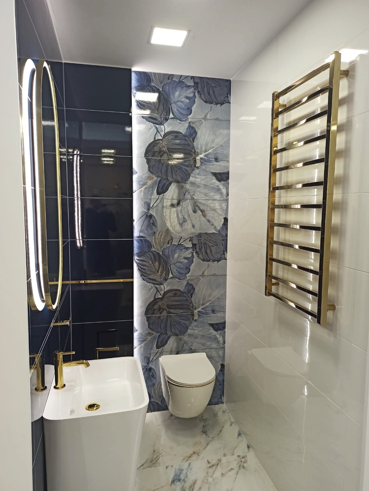 Nowoczesna łazienka z umywalką na postumencie, toaletą wiszącą i złotym grzejnikiem drabinkowym na białych płytkach, akcentowana czarnymi płytkami i płytkami z motywem liści.