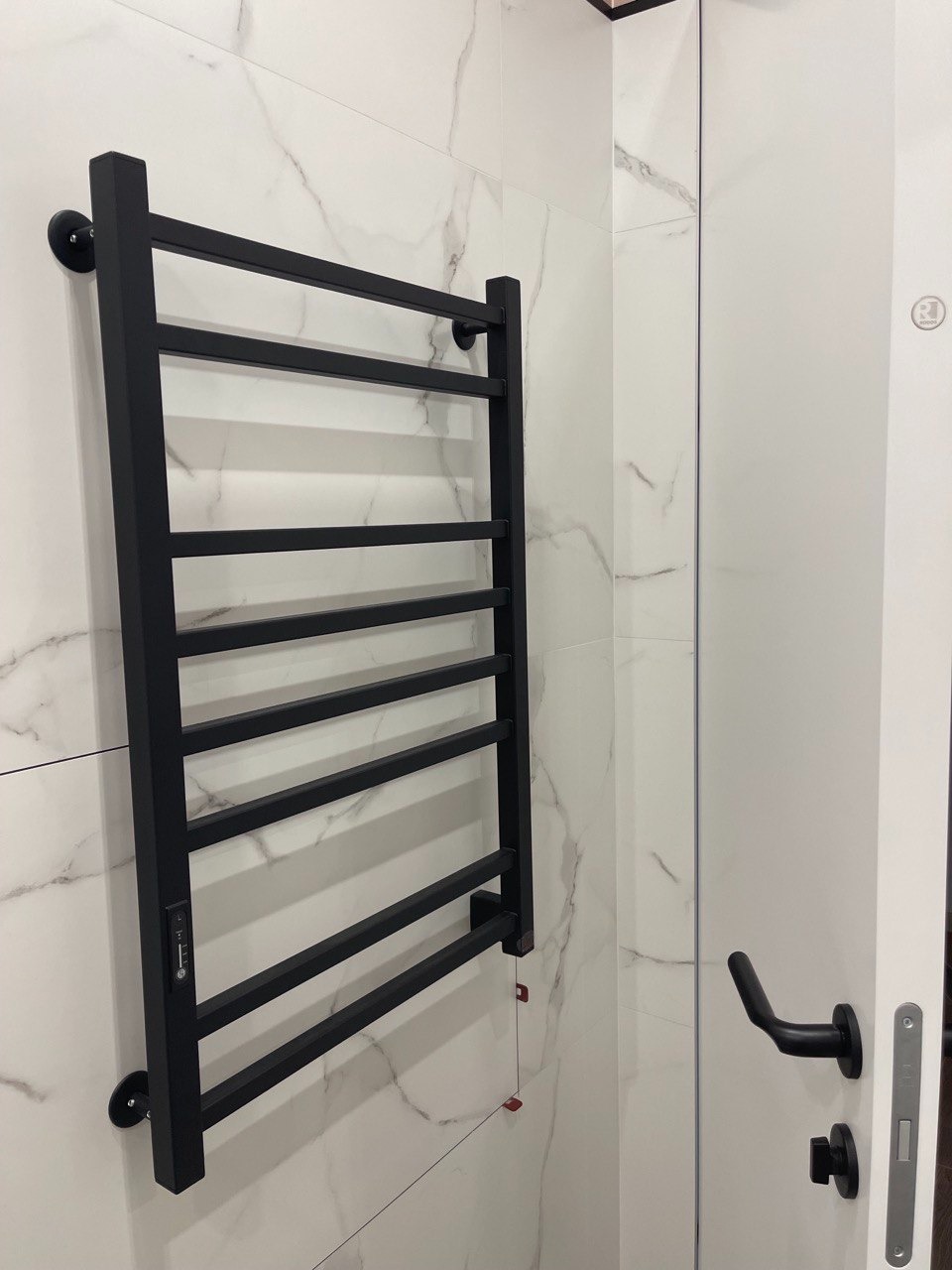 Czarny, elektryczny grzejnik drabinkowy łazienkowy z regulatorem temperatury, zamontowany na ścianie z białych płytek imitujących marmur. Widoczna czarna klamka drzwi.