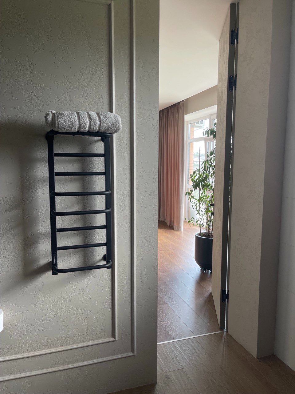 Czarny, elektryczny grzejnik drabinkowy z półką, zamontowany na teksturowanej ścianie w łazience z widocznym fragmentem drzwi i podłogi z drewna. Na grzejniku zwinięty biały ręcznik.