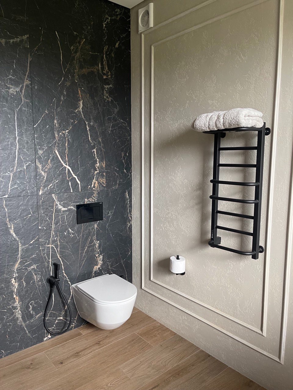 Nowoczesna łazienka z czarną marmurową ścianą, wiszącą toaletą z bidetką, drewnopodobną podłogą i czarnym, drabinkowym grzejnikiem z ręcznikiem na tle ściany w ramie.