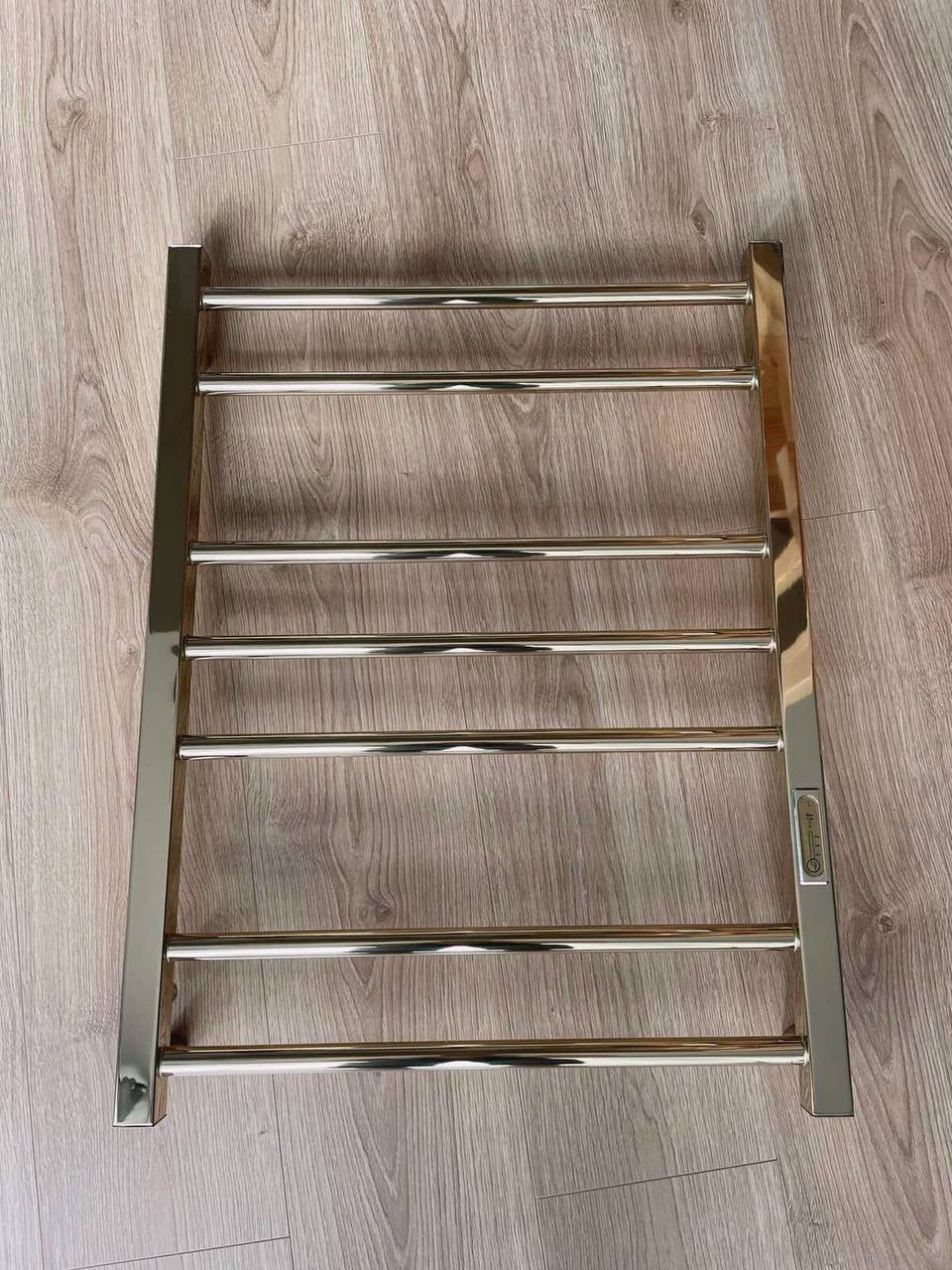 Grzejnik łazienkowy drabinkowy w kolorze złotym, leżący na drewnianej podłodze, widoczny z góry, z delikatnym odbiciem światła na powierzchni.