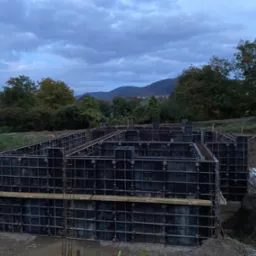 Szare, modułowe szalunki fundamentowe zbrojone stalą, przygotowane do zalania betonem, na tle zachmurzonego nieba i zieleni drzew oraz oddalonych wzg&oacute;rz.