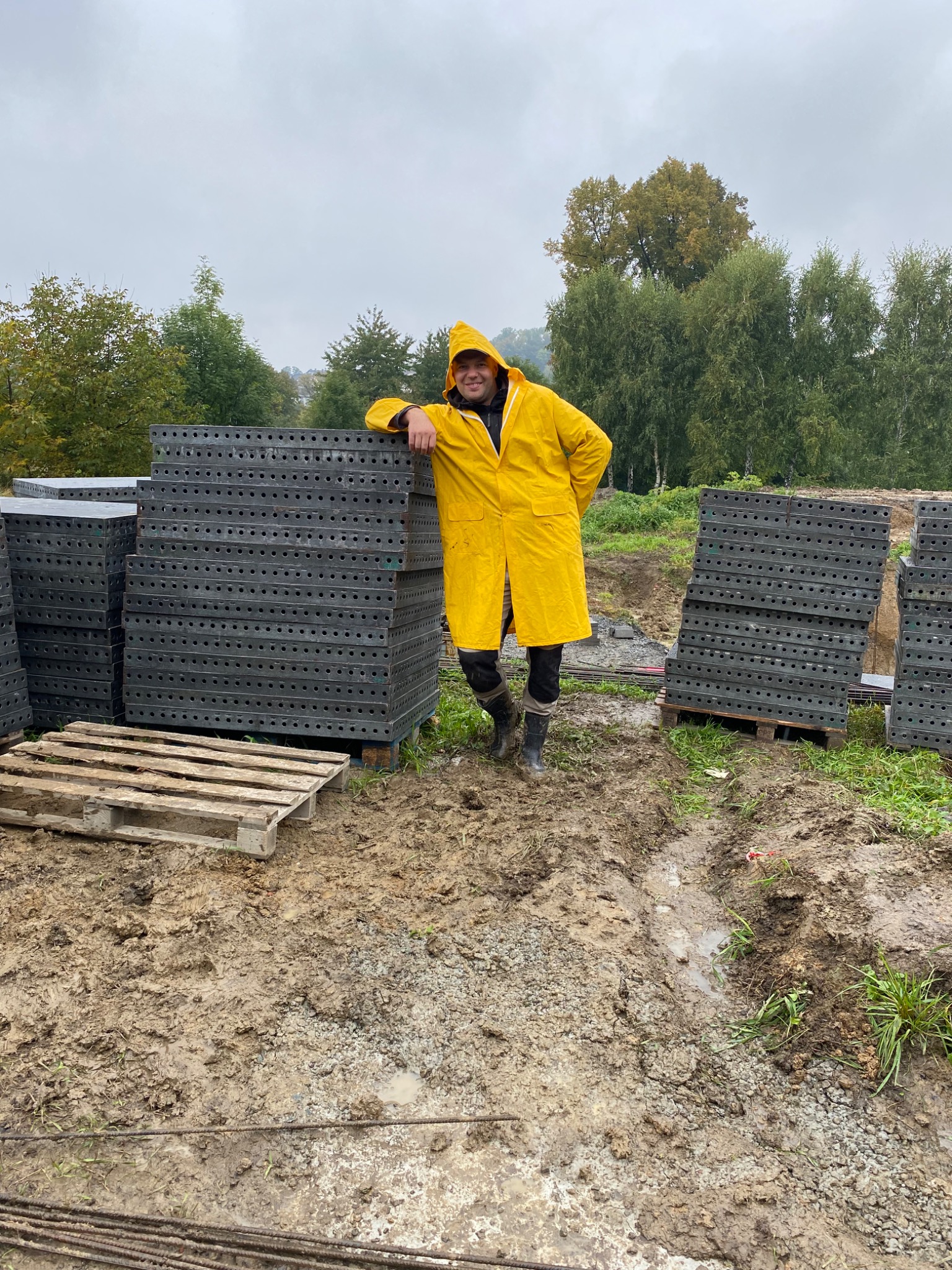 Osoba w żółtym płaszczu przeciwdeszczowym opiera się o stos szalunków budowlanych na placu budowy w pochmurny dzień.