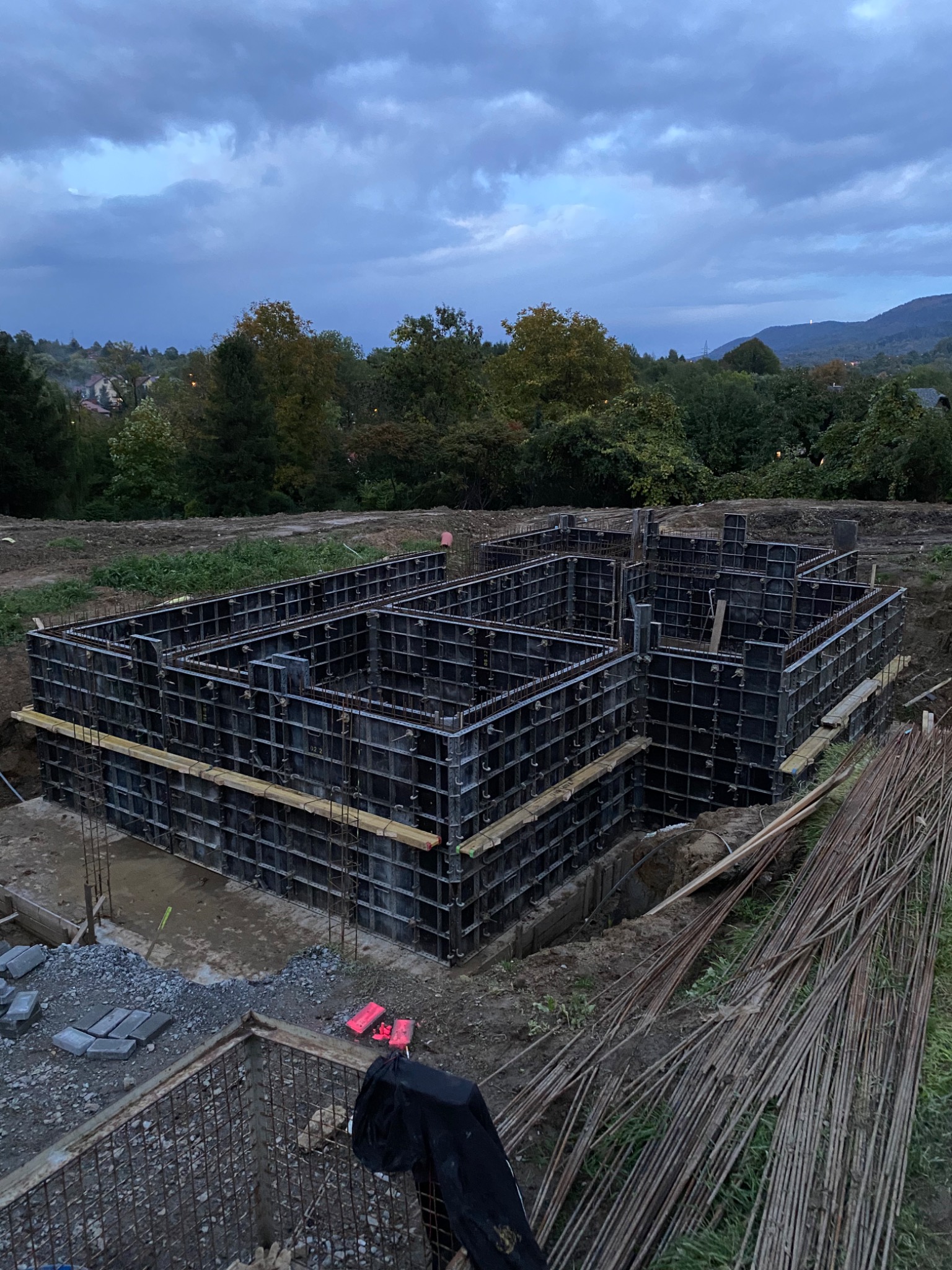 Szare, modułowe szalunki fundamentów domu jednorodzinnego, z widocznymi prętami zbrojeniowymi, na tle zachmurzonego nieba i zieleni drzew.