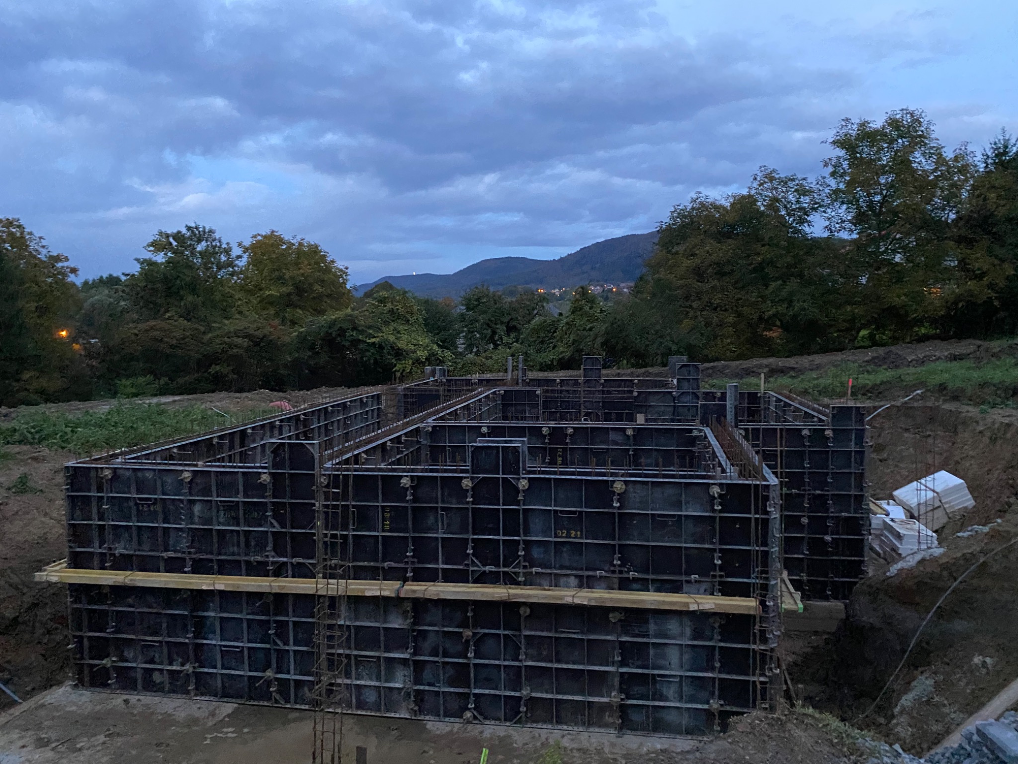 Szare, modułowe szalunki fundamentowe zbrojone stalą, przygotowane do zalania betonem, na tle zachmurzonego nieba i zieleni drzew oraz oddalonych wzgórz.