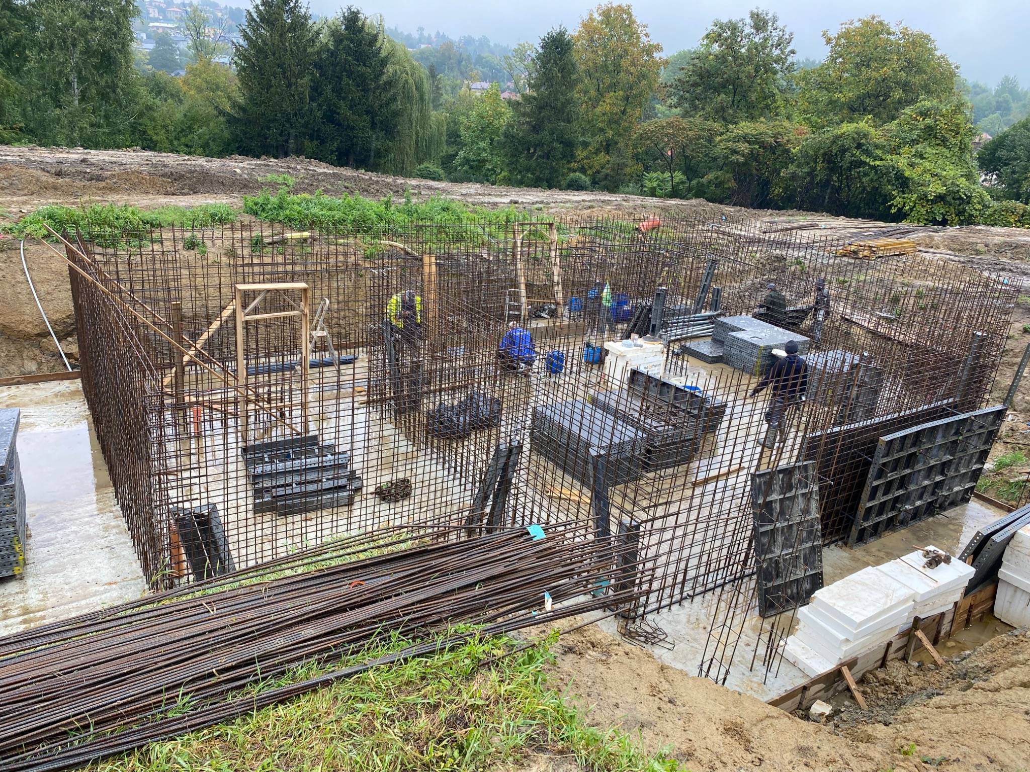 Zbrojenie fundamentów domu jednorodzinnego z widocznymi pracownikami w odblaskowych kamizelkach, szalunkami i stertą prętów zbrojeniowych na tle zieleni.