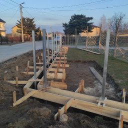 Patryk Mróz - Przygotowanie fundamentów pod ogrodzenie: drewniany szalunek z metalowymi słupkami w ziemi, widok na ulicę i domy w tle.