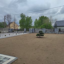 Patryk Mróz - Nowo założony ogród z ozdobnym drzewkiem bonsai na środku, otoczony kostką brukową i ogrodzeniem z elementów betonowych i metalowych przęseł, widoczne także lampki oświetleniowe wzdłuż ścieżki.