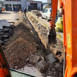 Patryk Mróz - Pomarańczowa koparka Kubota w trakcie prac ziemnych, wykopany wąski rów z ułożonymi niebieskimi rurami, obok sterta starych, szarych kostek brukowych, w tle budynek i zaparkowany biały samochód.