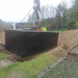 Patryk Mróz - Czarna, prefabrykowana skrzynia opuszczana dźwigiem do wykopu w ziemi, widoczny stos ziemi i fragment ogrodu, pochmurny dzień.