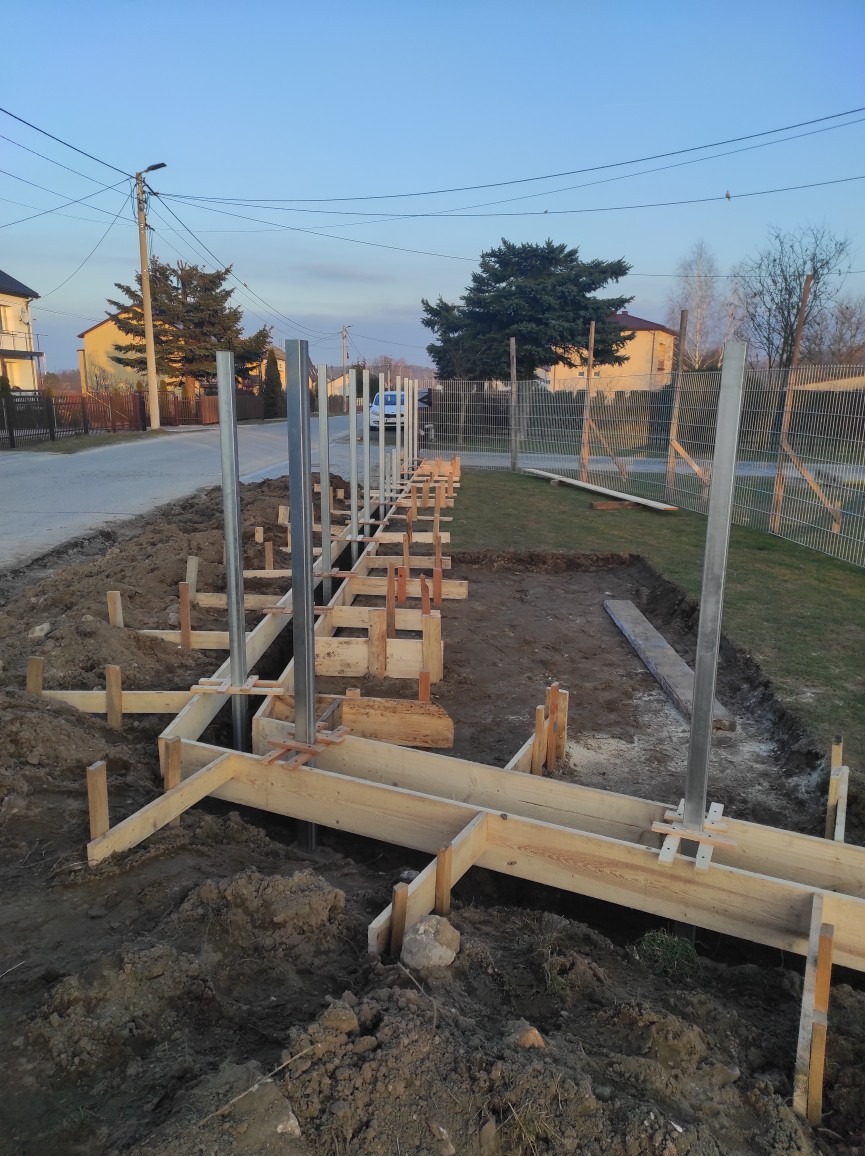 Przygotowanie fundamentów pod ogrodzenie: drewniany szalunek z metalowymi słupkami w ziemi, widok na ulicę i domy w tle.