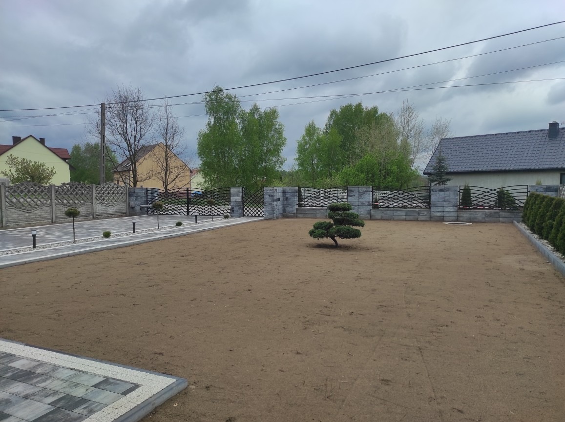 Nowo założony ogród z ozdobnym drzewkiem bonsai na środku, otoczony kostką brukową i ogrodzeniem z elementów betonowych i metalowych przęseł, widoczne także lampki oświetleniowe wzdłuż ścieżki.