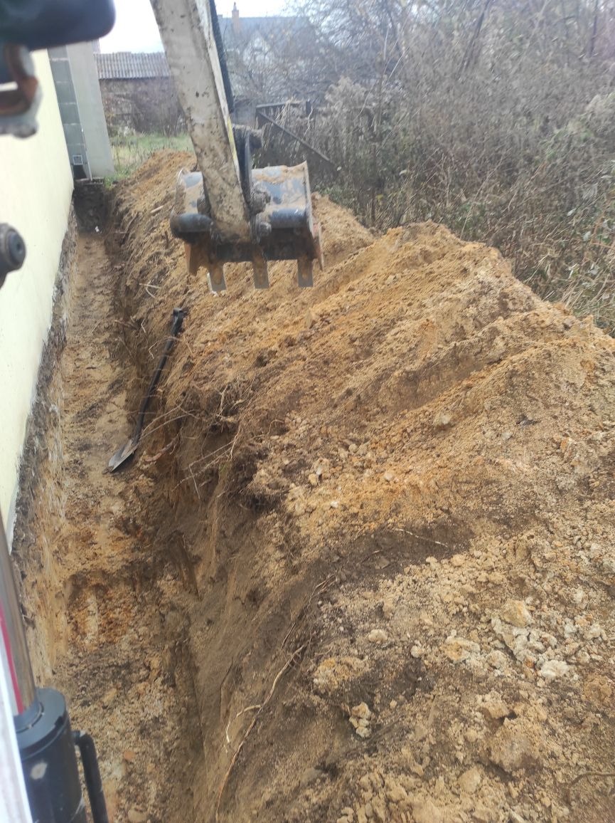Wąski wykop w ziemi wzdłuż ściany budynku, z widoczną łopatą w wykopie i łyżką koparki nad usypaną ziemią.