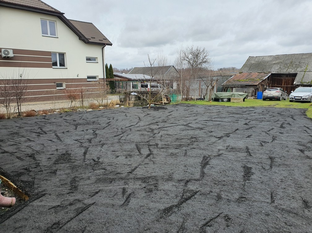 Przygotowany teren pod przyszłą kostkę brukową, wyrównany i pokryty czarnym materiałem, w tle dom jednorodzinny i budynki gospodarcze w pochmurny dzień.