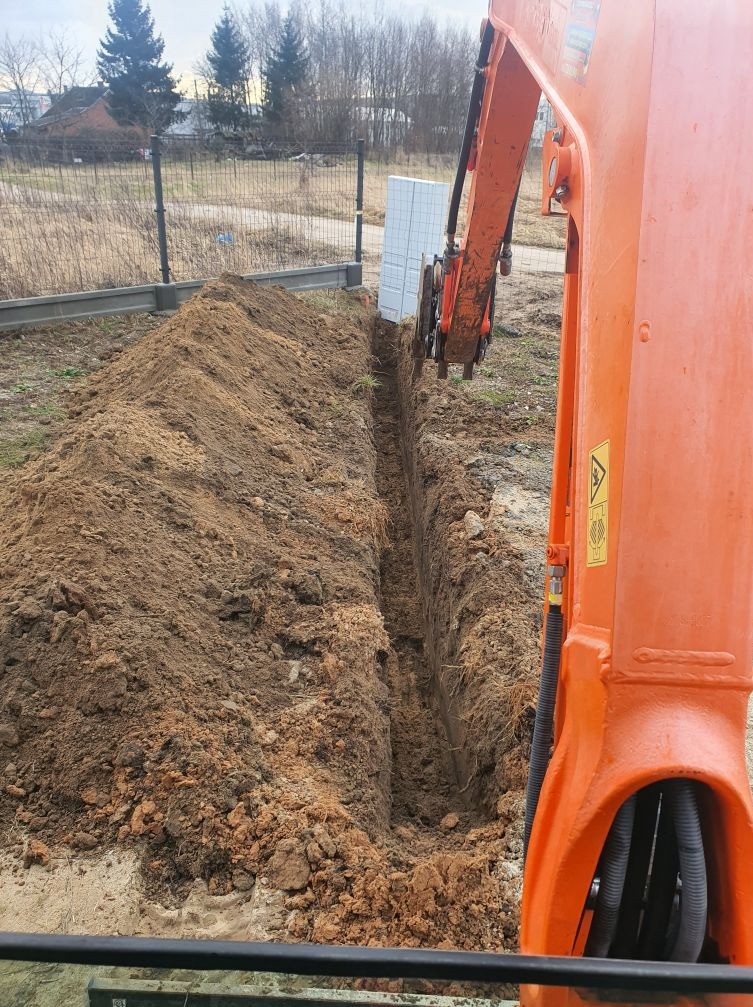 Pomarańczowa koparka w trakcie wykonywania wąskiego wykopu w ziemi, obok widoczny stos białych bloczków, w tle ogrodzenie panelowe i zabudowania.