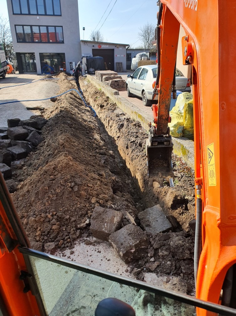 Pomarańczowa koparka Kubota w trakcie prac ziemnych, wykopany wąski rów z ułożonymi niebieskimi rurami, obok sterta starych, szarych kostek brukowych, w tle budynek i zaparkowany biały samochód.