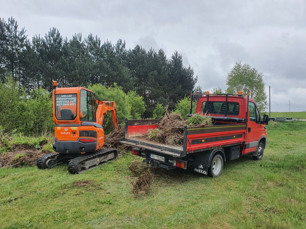 Pomarańczowa minikoparka Kubota i czerwony samochód dostawczy Iveco z wywrotką, załadowany korzeniami i fragmentami roślin, na zielonym trawiastym terenie, w tle las iglasty.