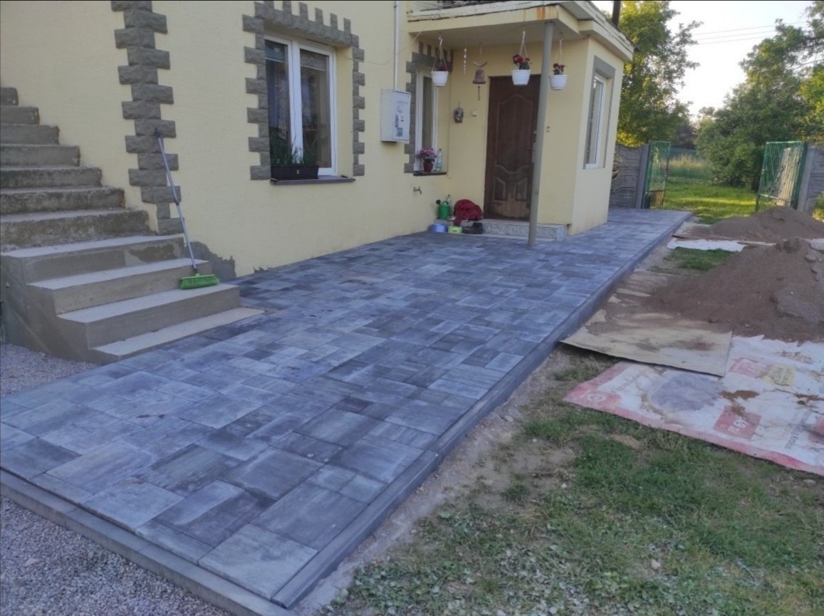 Nowo ułożona, szara kostka brukowa prowadząca od schodów wejściowych do domu; widoczny fragment trawnika i pryzma piasku.