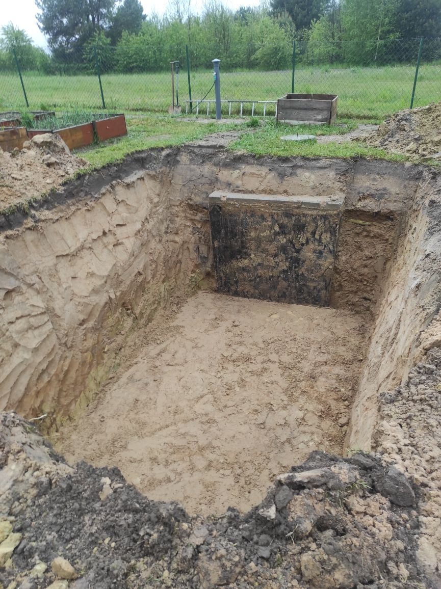 Głęboki wykop w ziemi z widocznym betonowym elementem konstrukcyjnym w jednej ze ścian, na górze widoczna trawa i ogrodzenie z siatki.