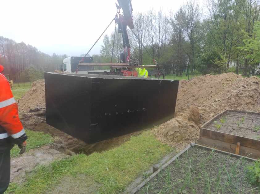 Czarna, prefabrykowana skrzynia opuszczana dźwigiem do wykopu w ziemi, widoczny stos ziemi i fragment ogrodu, pochmurny dzień.